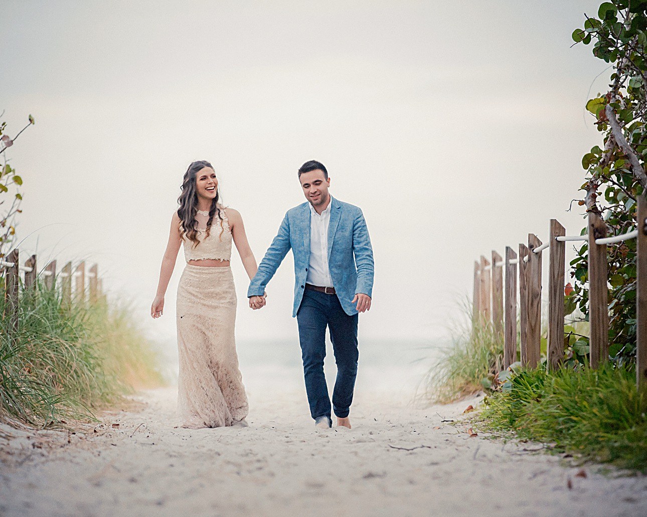 Una pareja camina de la mano por un sendero de arena hacia la playa, riendo
