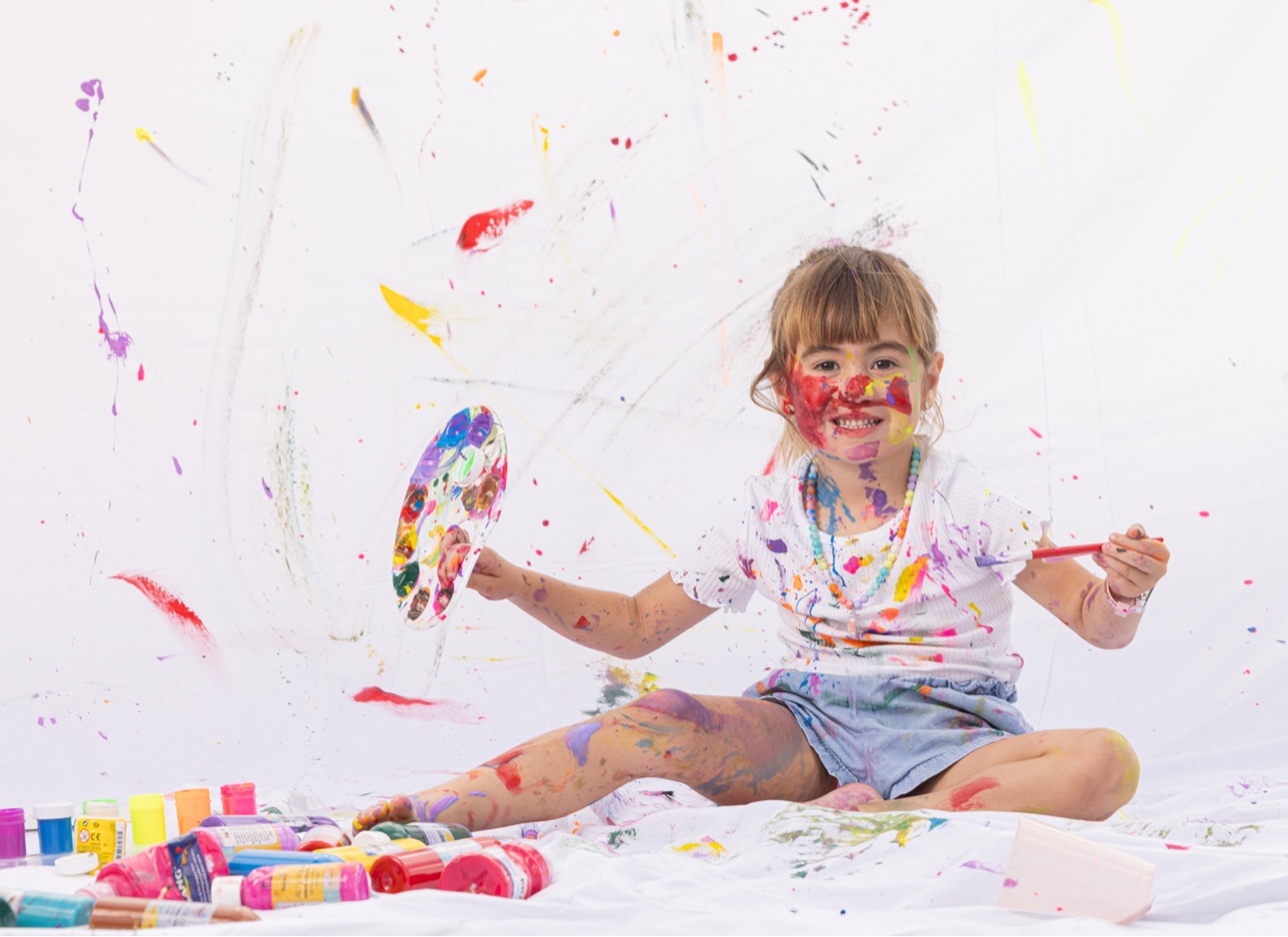 Niña sentada en el piso con paleta y pincel, rodeada de pinturas de colores, en su Paint Day Party